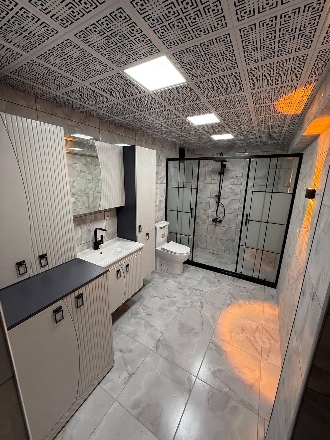 Siyah çerçeveli duş alanı ve krem rengi dolaplarla çevrili bir banyo