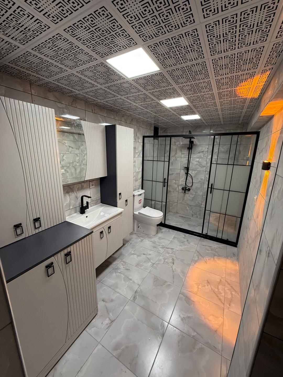 Siyah çerçeveli duş alanı ve krem rengi dolaplarla çevrili bir banyo