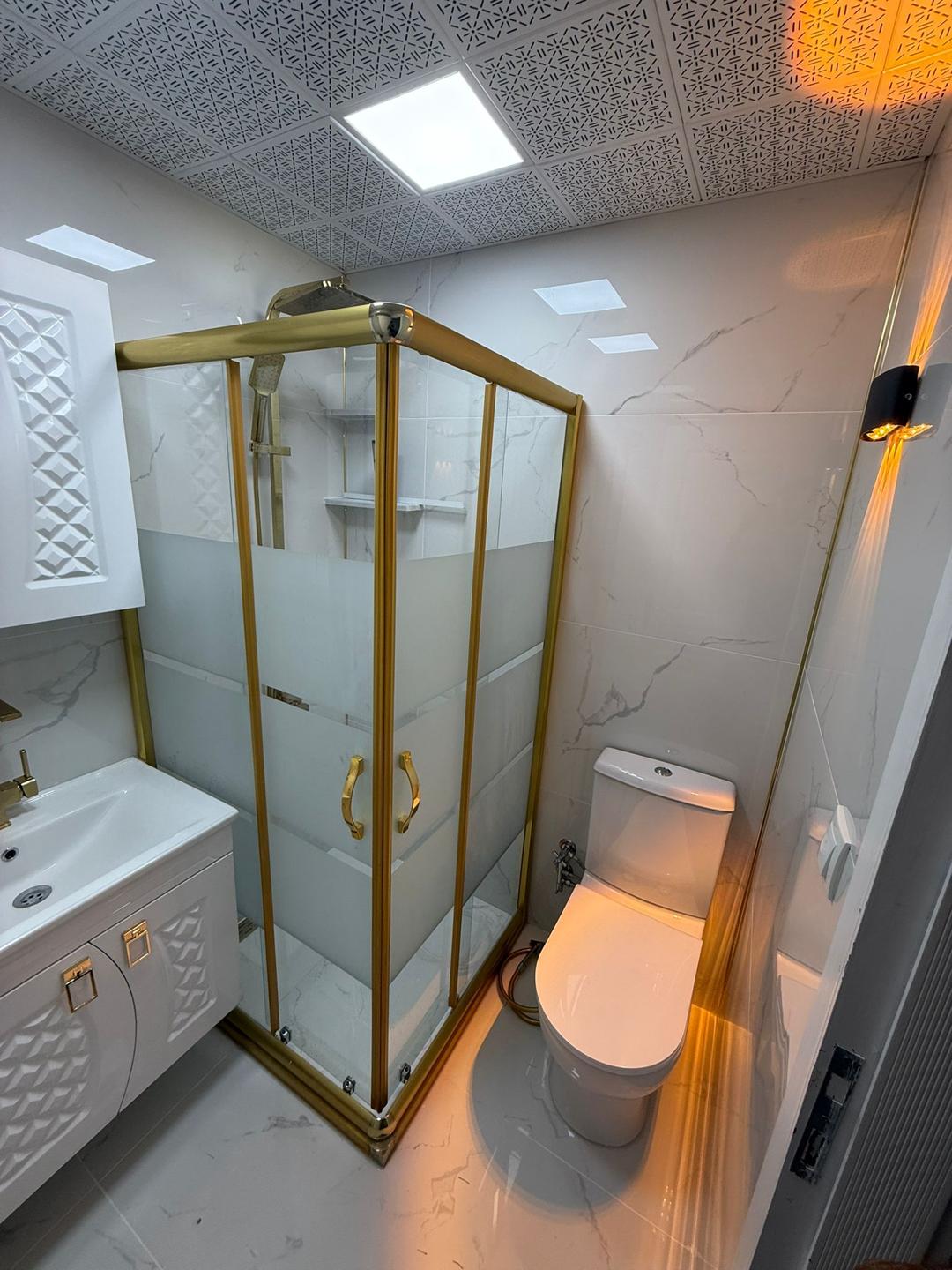 Metalik altın rengi çerçeveli bir duşakabin ve beyaz dolaplarla donatılmış bir banyo